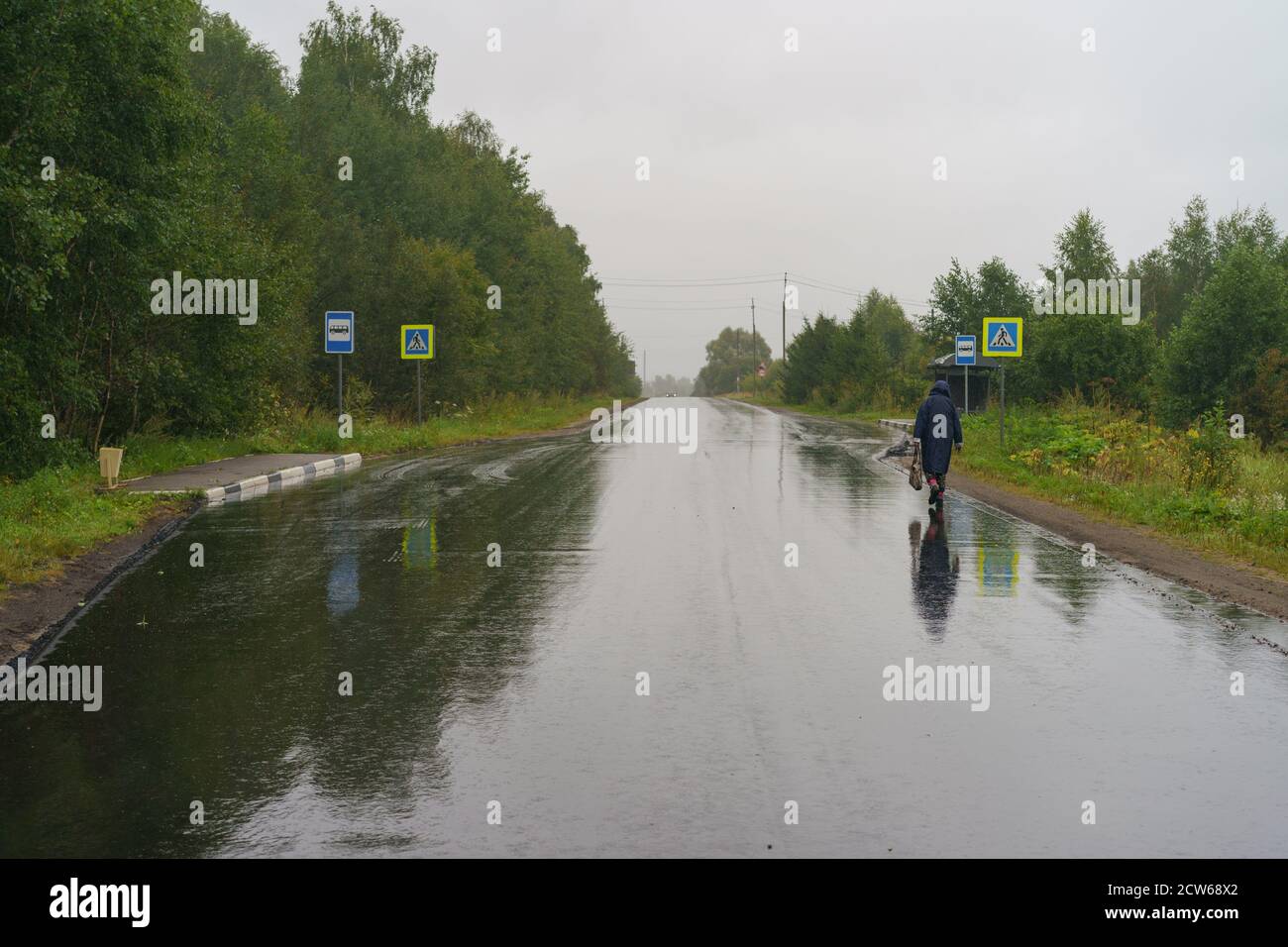 Der Herbst kommt in die russische Landschaft. Nasse Straße tagsüber. Kalte Dusche. Einsame Frau zog Regenmantel an, um zur Bushaltestelle zu laufen. Inlandstourismus Stockfoto