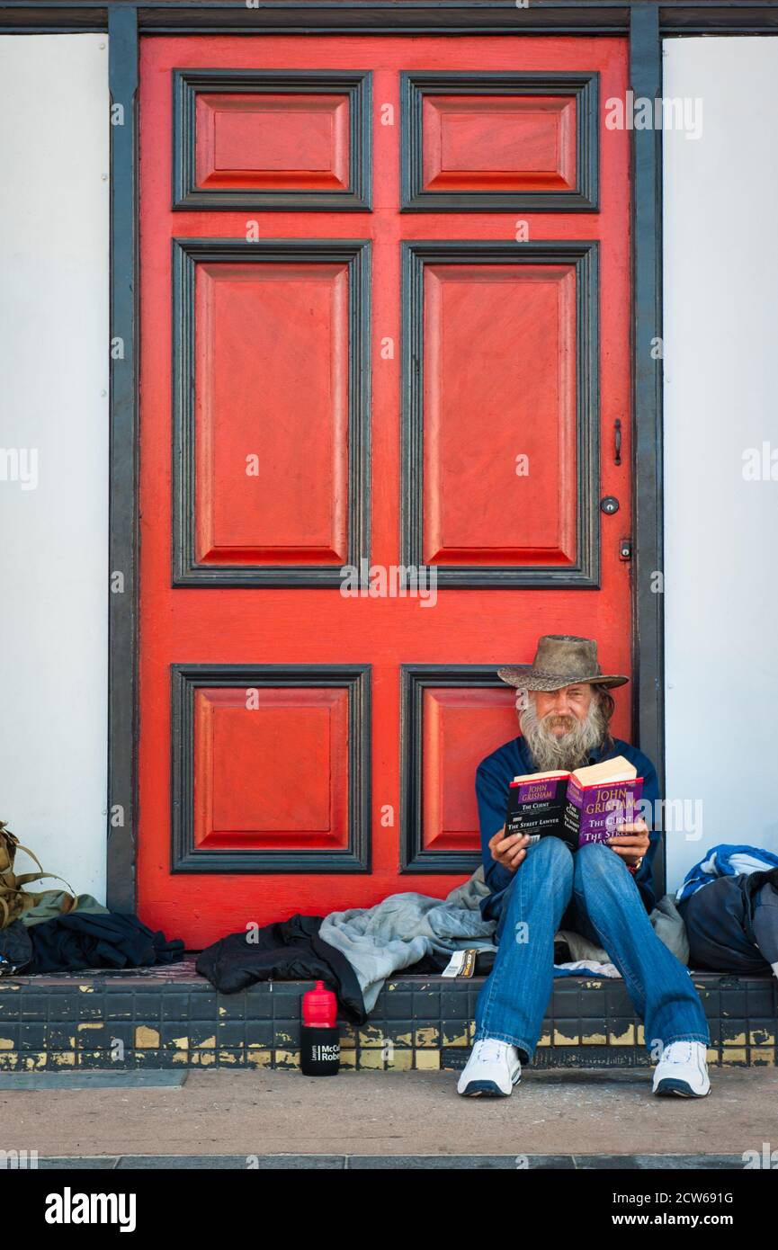 Ein alter Mann sitzt vor einer rot-schwarzen Tür und liest einen Roman, während er eine Pause von seiner Solo-Reise durch Townsville in QLD, Australien macht. Stockfoto