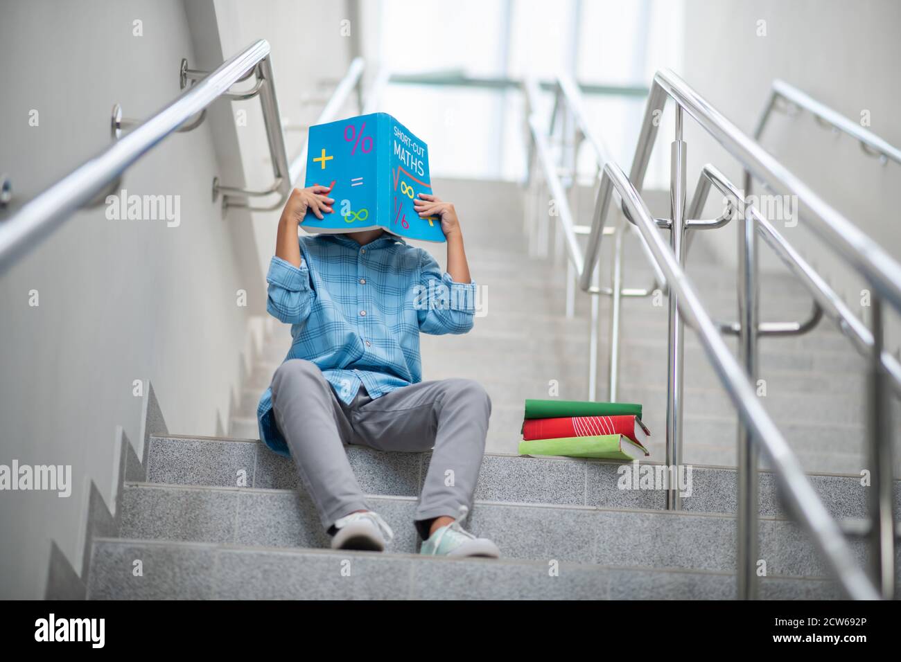 Schuljunge sitzt auf der Treppe, bedeckt sein Gesicht mit Mathematik Buch Stockfoto