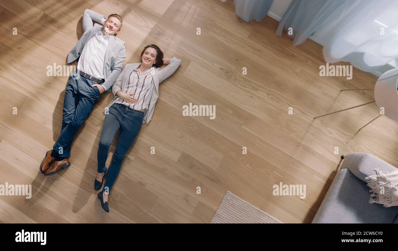 Das junge Paar liegt auf einem Holzboden in einer Wohnung. Sie sind glücklich, lächeln und lachen. Gemütliches Wohnzimmer mit moderner Einrichtung, grauem Sofa und Stockfoto