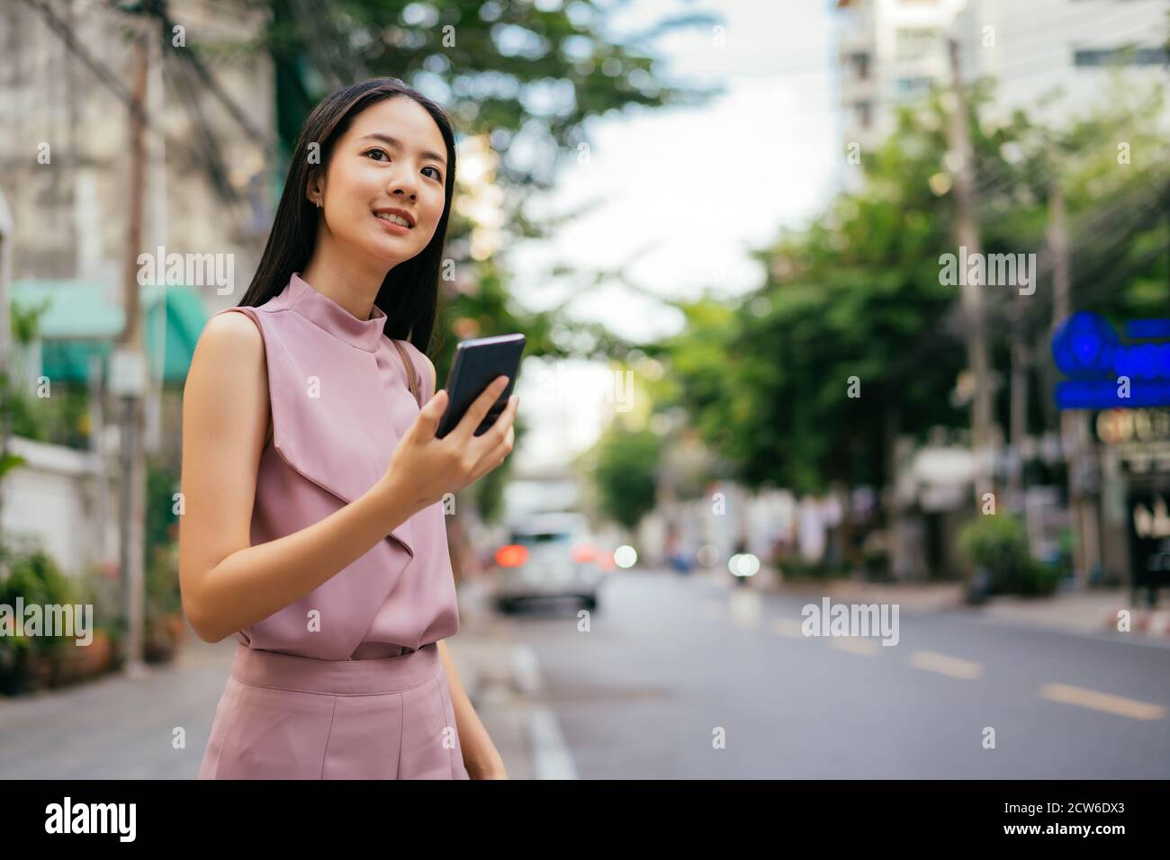 Glückliche asiatische Geschäftsfrau am Straßenrand hält Smartphone, mit App, um ein Taxi zu buchen, Kommunikation, Verbindung, unterwegs Stockfoto