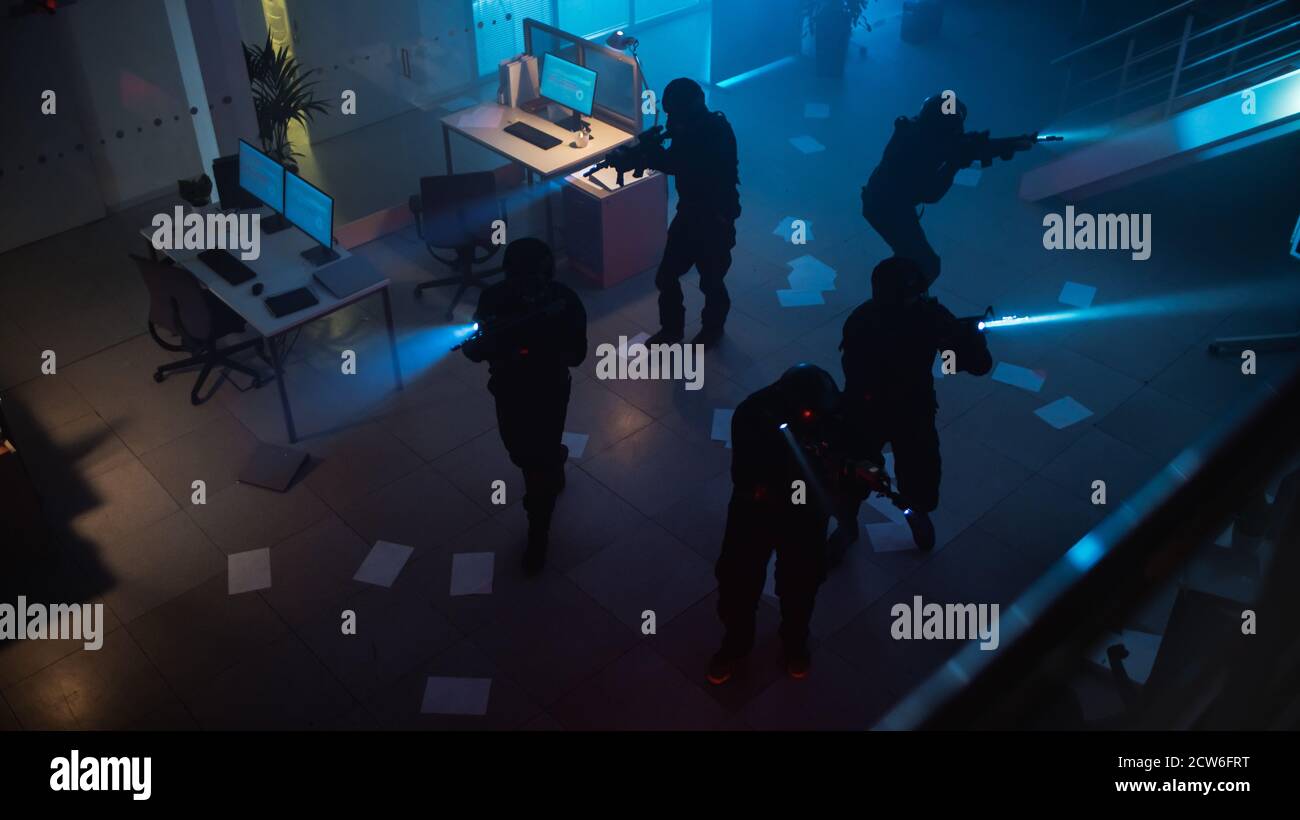 Maskierte Truppe bewaffneter SWAT-Polizisten stürmt ein finsteres beschlagnahmtes Bürogebäude mit Schreibtischen und Computern. Soldaten mit Gewehren und Taschenlampen bewegen sich Stockfoto