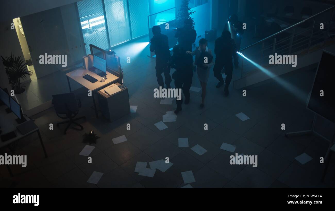 Maskierte Truppe bewaffneter SWAT-Polizeibeamter rettet eine weibliche Geisel in einem dunklen beschlagnahmten Bürogebäude mit Schreibtischen und Computern. Soldaten mit Gewehren und Stockfoto