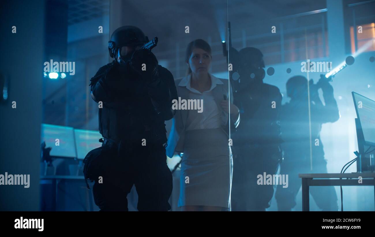 Maskierte Truppe bewaffneter SWAT-Polizeibeamter rettet eine weibliche Geisel in einem dunklen beschlagnahmten Bürogebäude mit Schreibtischen und Computern. Soldaten mit Gewehren und Stockfoto