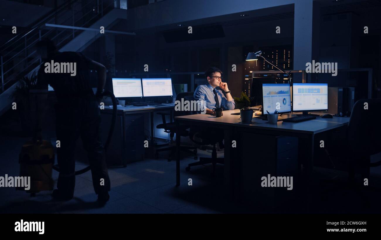 Arbeiten spät im Büro: Geschäftsmann nutzt Desktop-Computer, Analyse, Verwendung von Dokumenten, Probleme lösen, wichtige Projekt abschließen. Gewissenhaft Stockfoto