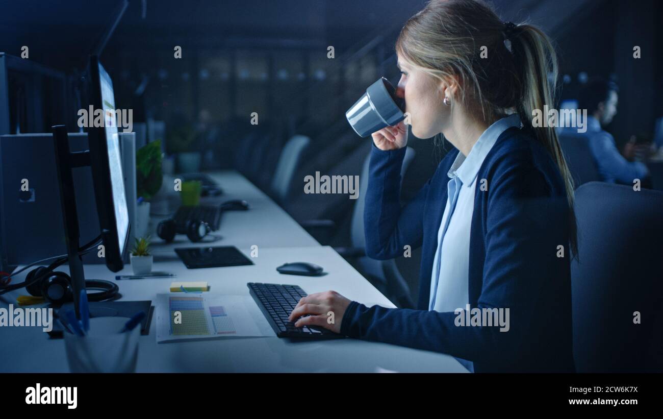 Arbeiten spät in der Nacht im Büro: Geschäftsfrau mit Desktop-Computer und Kaffee trinken, analysieren, mit Dokumenten, Probleme lösen, Finishing Stockfoto