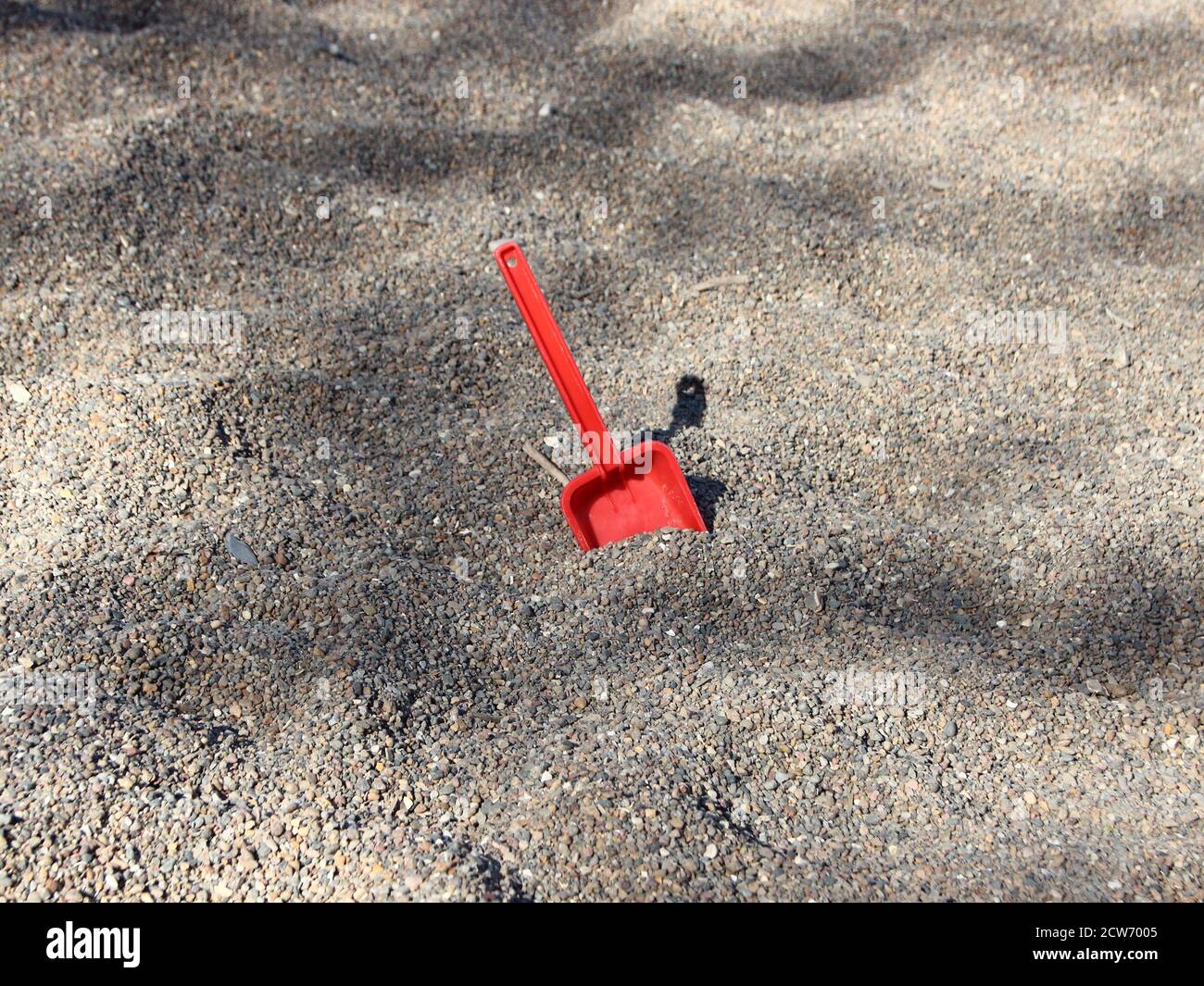 Eine einzelne rote Schaufel, die in einer Sandkiste des Kindergartens zurückgelassen wurde, die rote Schaufel kontrastiert den grauen Kies gewaltig. Stockfoto