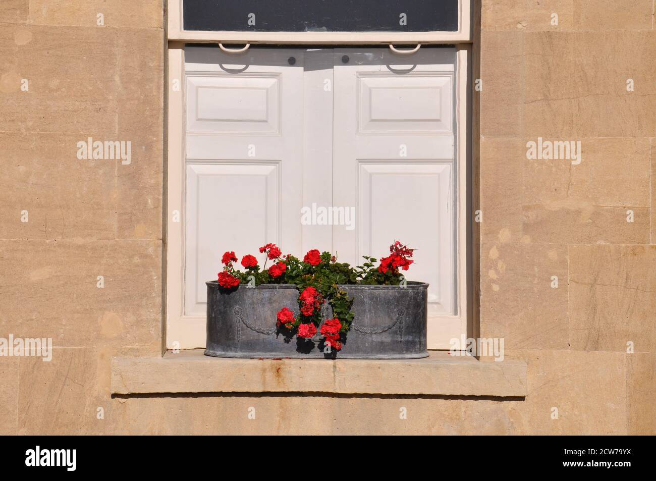 Fensterkasten aus Eisengepflanzer mit roten Geranien auf einer Sandsteinfensterschwelle vor geschlossenen weißen Fensterläden aus Holz. Stockfoto