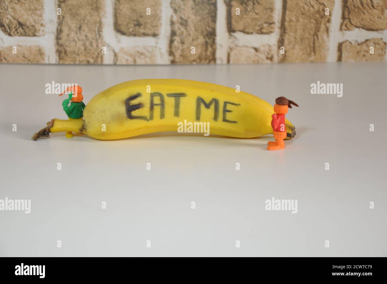 Banane. Obst mit Inschrift, ISS mich, mit Miniatur-Plastikspielzeug, das auf Obst schaut, weißer Sockel, Backsteinwand-Hintergrund, in Konzeptfoto, Brasilien, Stockfoto