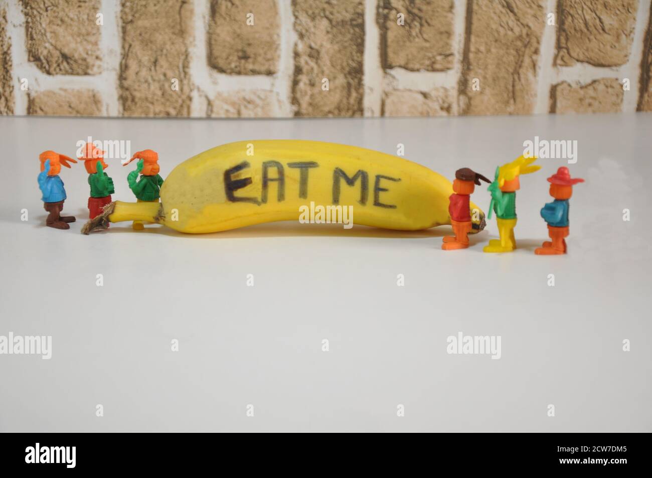 Banane. Obst mit Inschrift, ISS mich, mit Miniatur-Plastikspielzeug, das auf Obst schaut, weißer Sockel, Backsteinwand-Hintergrund, in Konzeptfoto, Brasilien, Stockfoto