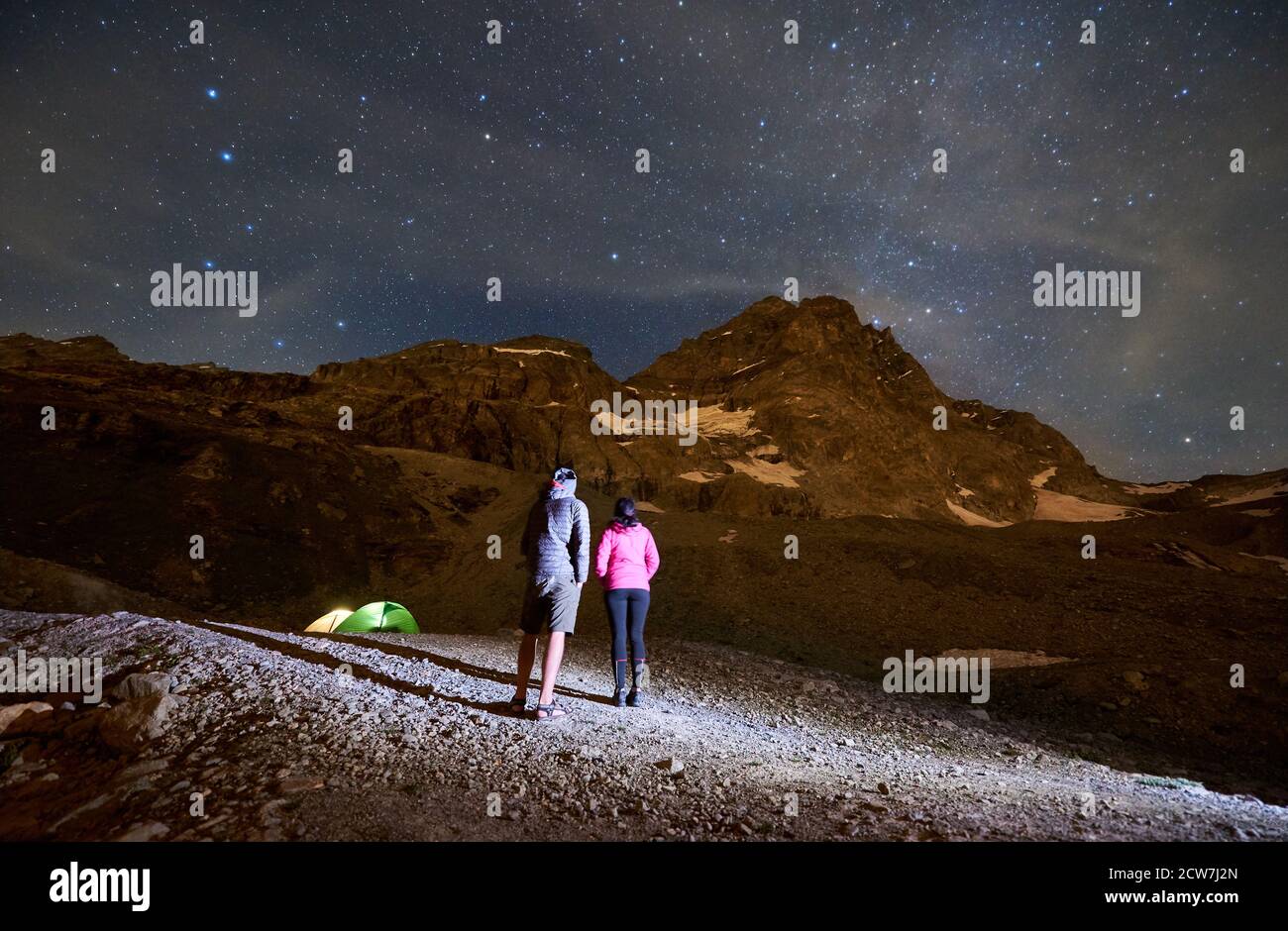 Rückansicht der Wanderer bewundern Blick auf majestätische Berge unter Nachthimmel mit Starts. Frau und Mann stehen auf einem felsigen Hügel und genießen die nächtliche Berglandschaft. Konzept von Reisen, Wandern, Nacht. Stockfoto