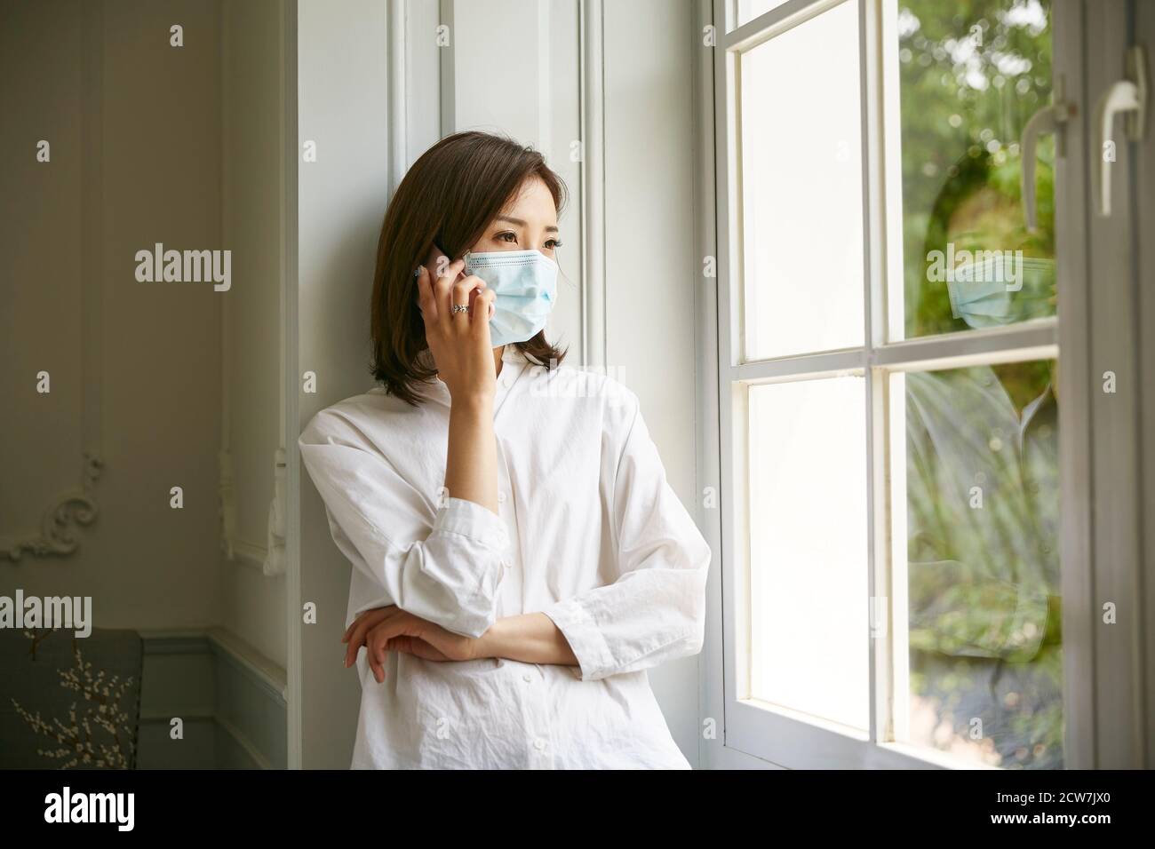 Junge asiatische Frau in Isolation trägt Maske am Fenster stehen Telefonieren Stockfoto
