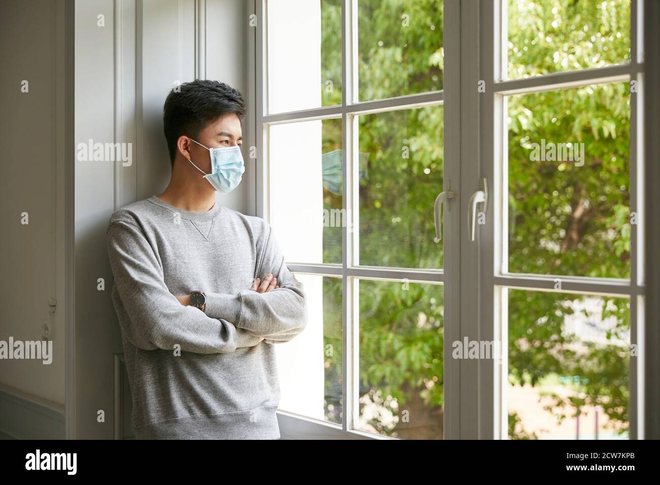 Junger asiatischer Mann, der am Fenster zu Hause mit Maske steht Arme gekreuzt Stockfoto