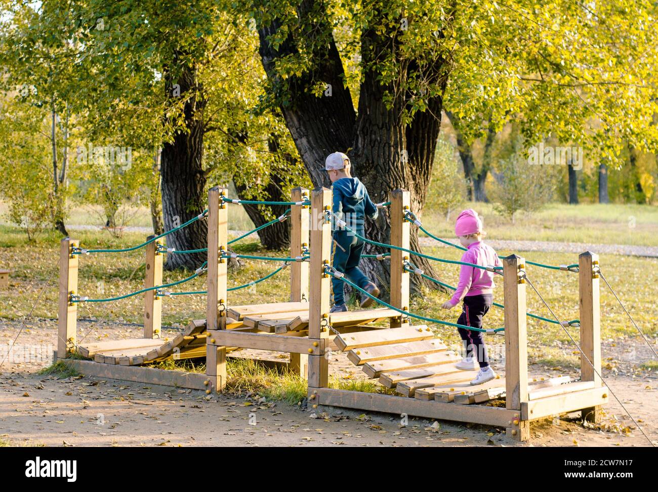 Hölzerne Spielbrücke für Kinder, im Park unter den Bäumen gelegen. Spielplätze im Freien. Eine Brücke aus Holzbohlen mit einem Seilgeländer, wo s Stockfoto