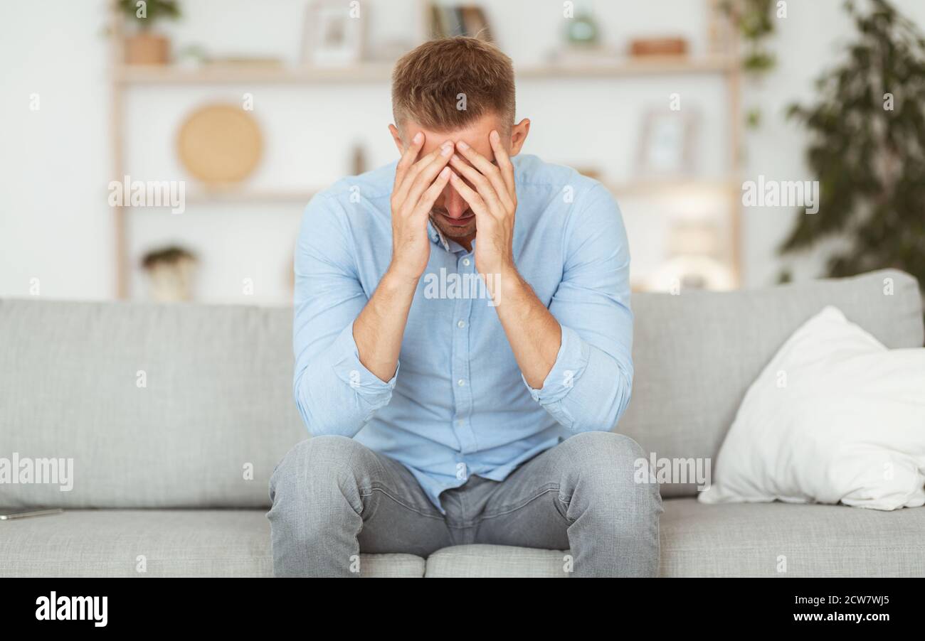 Deprimierter Kerl, der auf der Couch sitzt und das Gesicht mit den Händen bedeckt Stockfoto