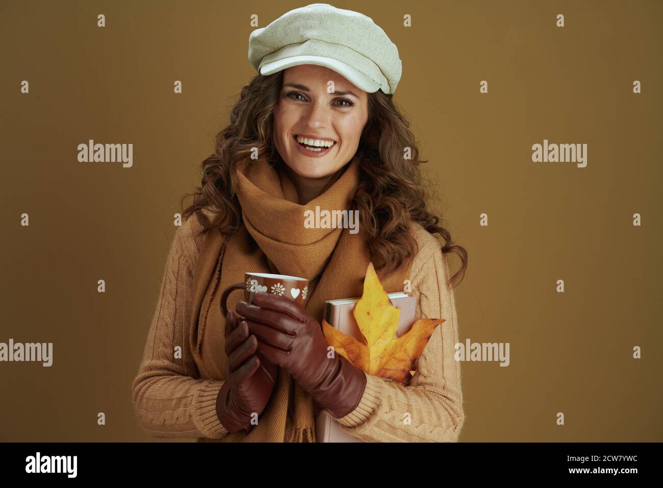 Hallo september. Portrait einer glücklichen modernen Frau im Schal mit gelben Herbst Ahornblatt, Buch, Lederhandschuhe und Tasse vor beigem Hintergrund. Stockfoto