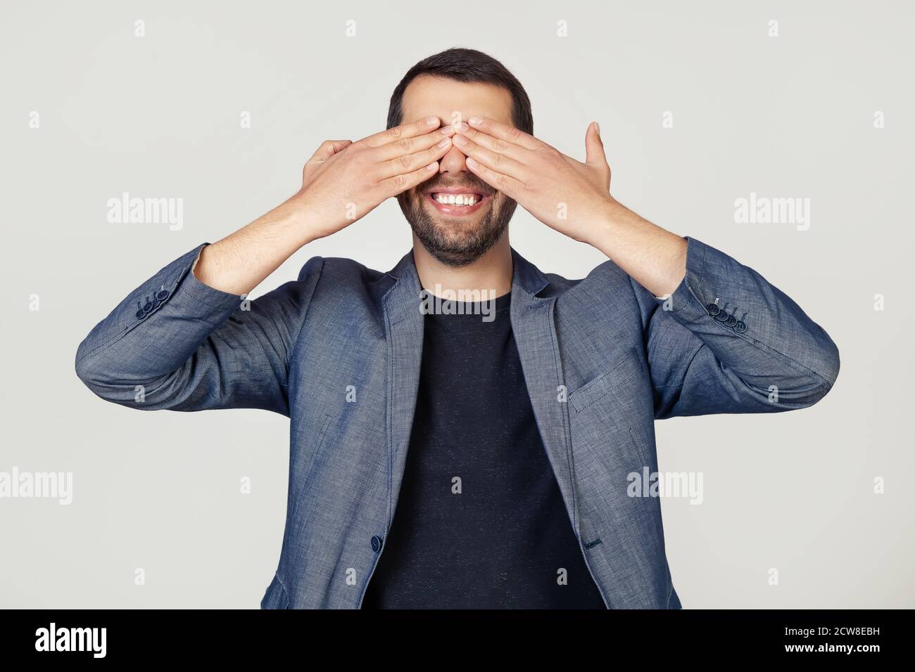 Junger Geschäftsmann Mann mit einem Bart in einer Jacke bedeckt seine Augen mit seinen Händen lächelnd fröhlich und lustig. Blindes Konzept. Porträt eines Mannes auf grauem Hintergrund. Stockfoto