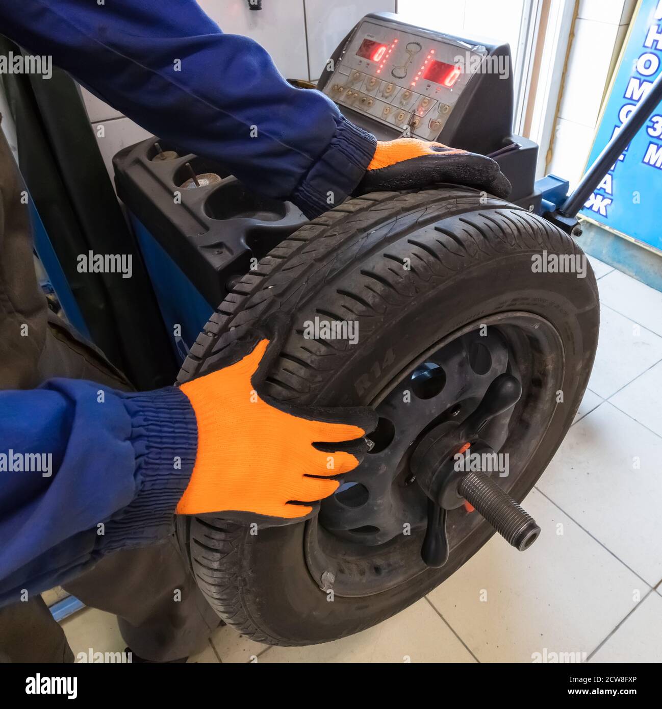 Mechaniker arbeitet mit Radauswuchtmaschine bei Reifenservice, Nahaufnahme. Mechaniker trägt orangefarbene Handschuhe und blaues Outfit. Stockfoto