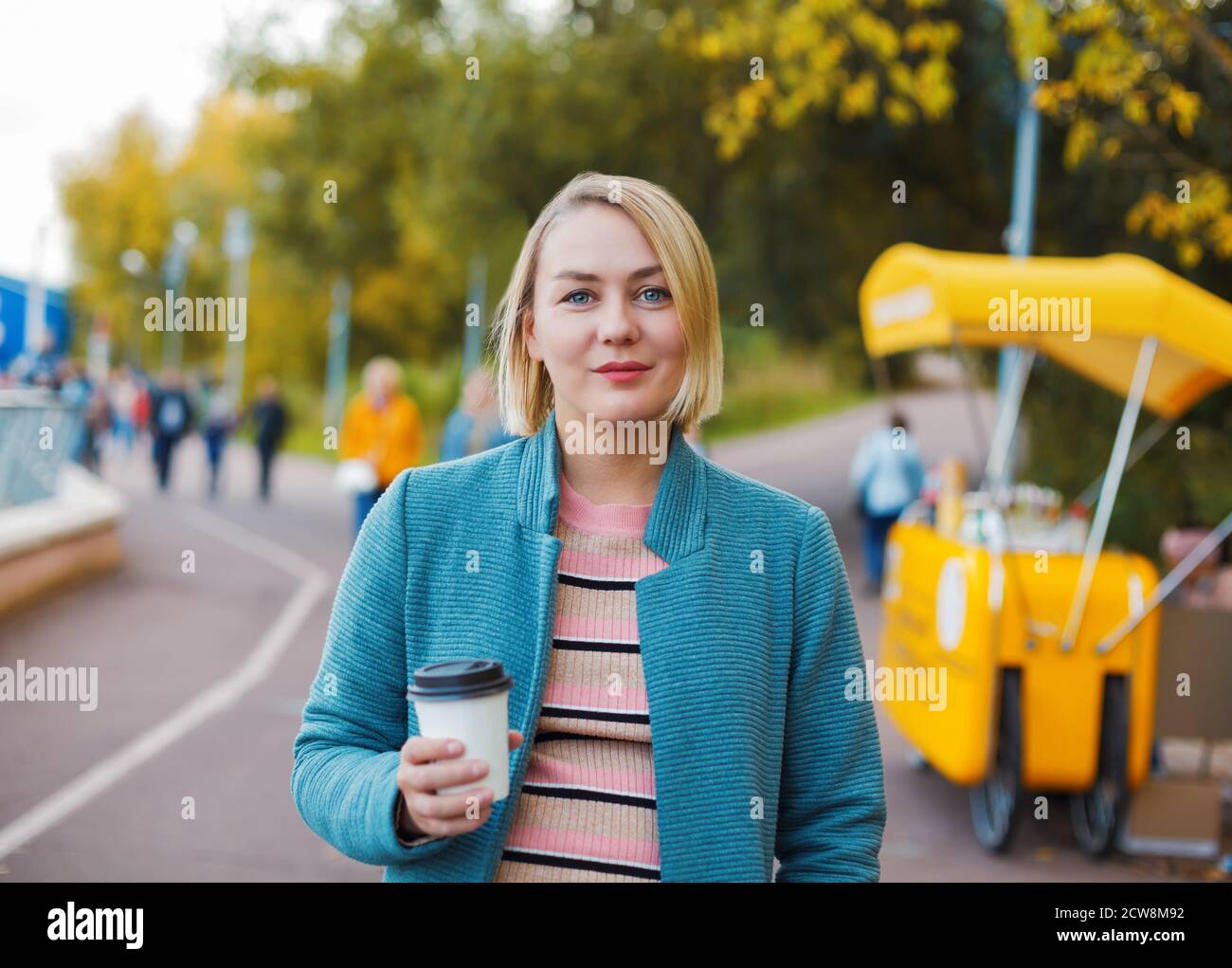 Lächelnde blonde kaukasische Frau mit Kaffeetasse im Herbst Park. Outdoor-Porträt während des indischen Sommers. Stockfoto