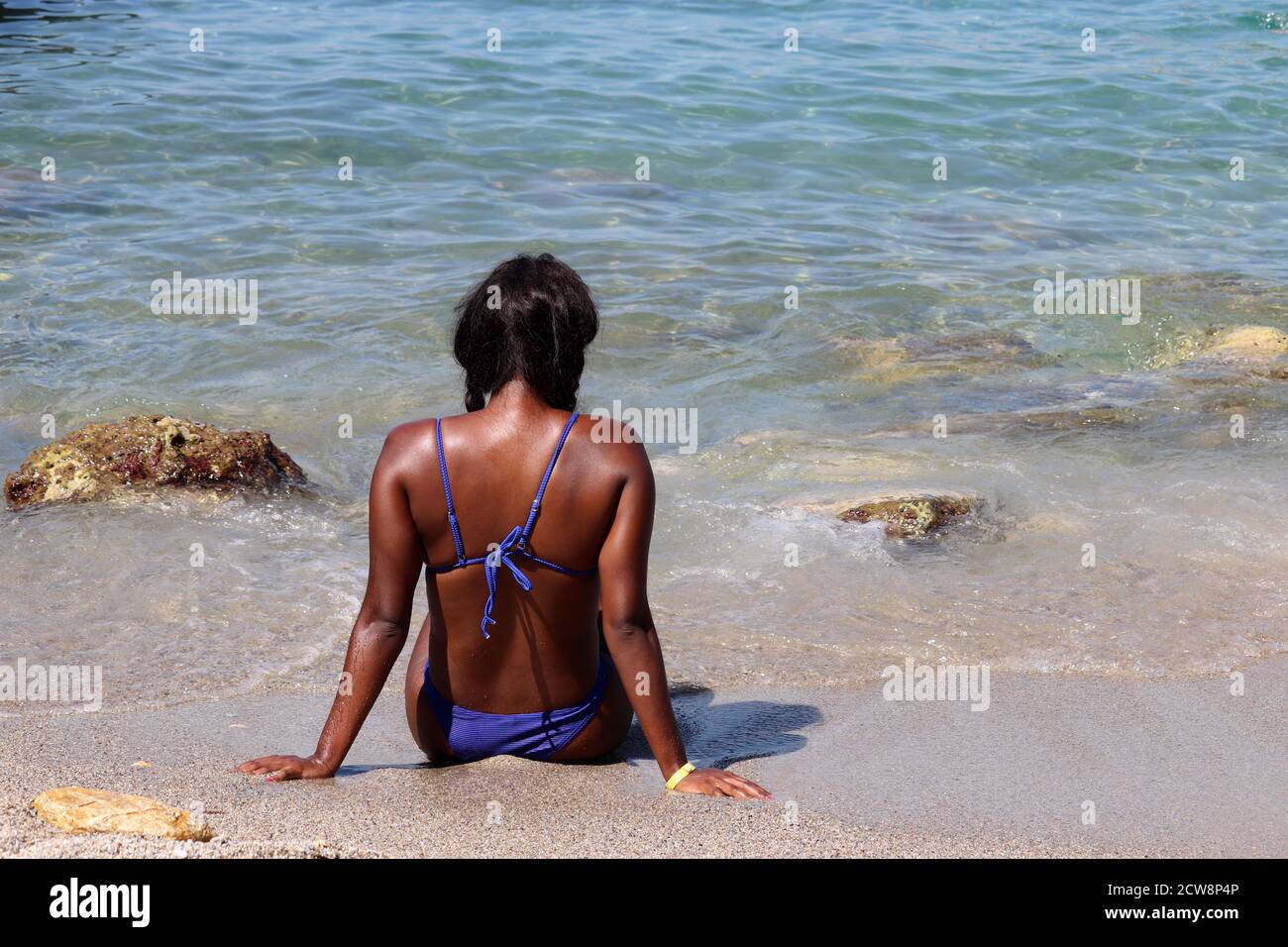 Schwarze Frau in blauen Bikini Sonnenbaden an einem Sandstrand sitzen auf Wellen Hintergrund. Urlaub, Entspannung und Freizeit am Meer Stockfoto