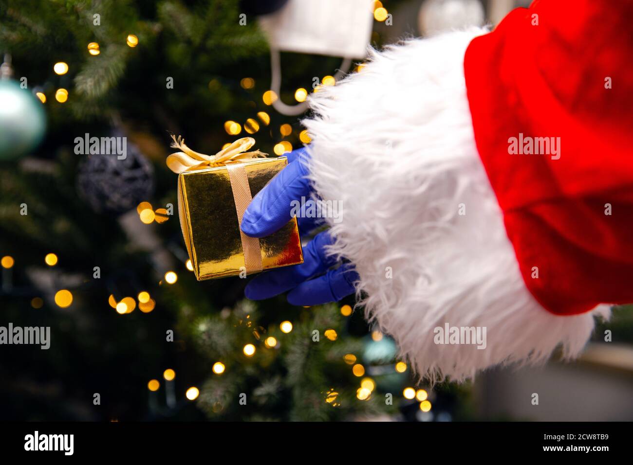 Weihnachtsmann mit Handschuhen und Gesichtsmaske für Coronavirus am Weihnachtsbaum, mit goldenem Geschenk, Covid-19 und Weihnachts-Sicherheitskonzept Stockfoto