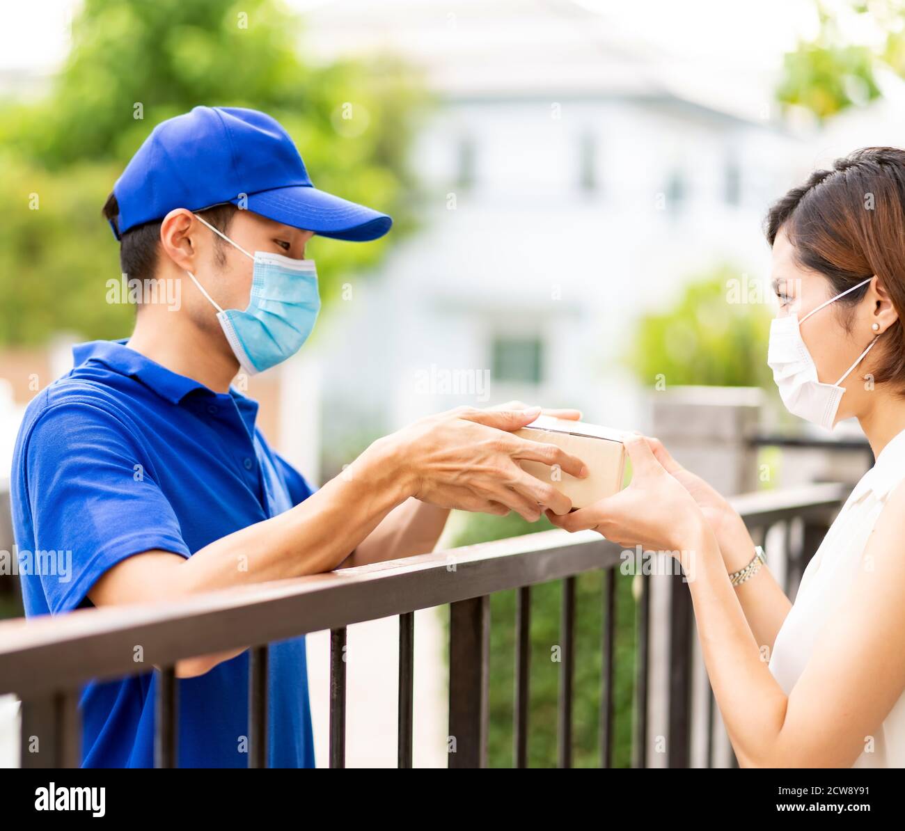Asian liefern Mann in blauem Hemd Handhabung Paket und geben an junge Frau Kunden zu Hause. Sie tragen Gesichtsmaske, um Coronavirus COVID-19 spreadi zu reduzieren Stockfoto