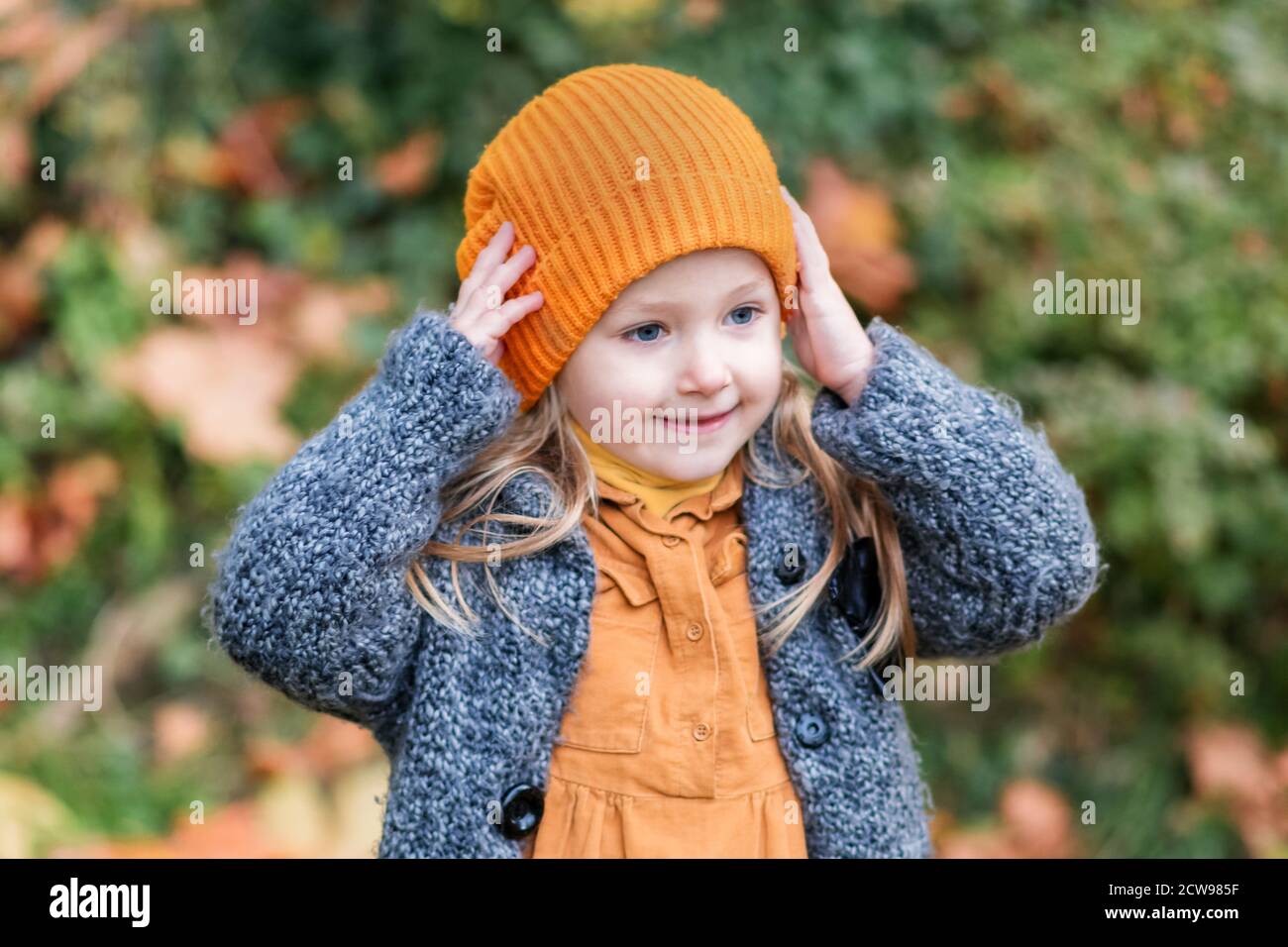 Ein kleines Mädchen in einem orangen Hut und einem grauen Mantel hält im Herbst in einem Park. Stockfoto
