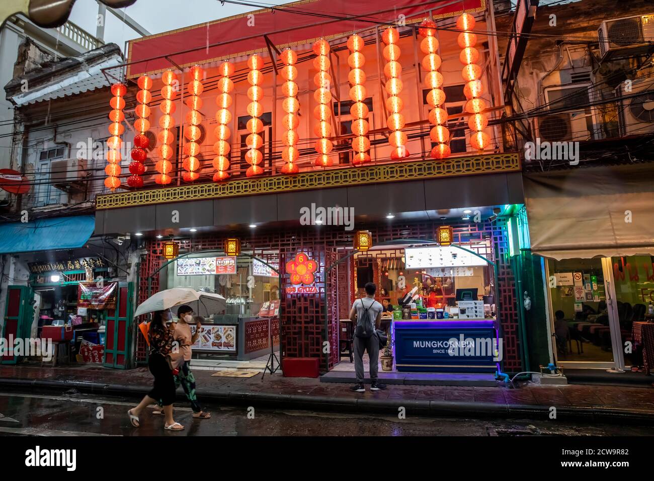 Ein Paar geht die Straße unter Sonnenschirmen entlang, während ein anderer Mann in ein Restaurant in Bangkoks Chinatown schaut. Stockfoto