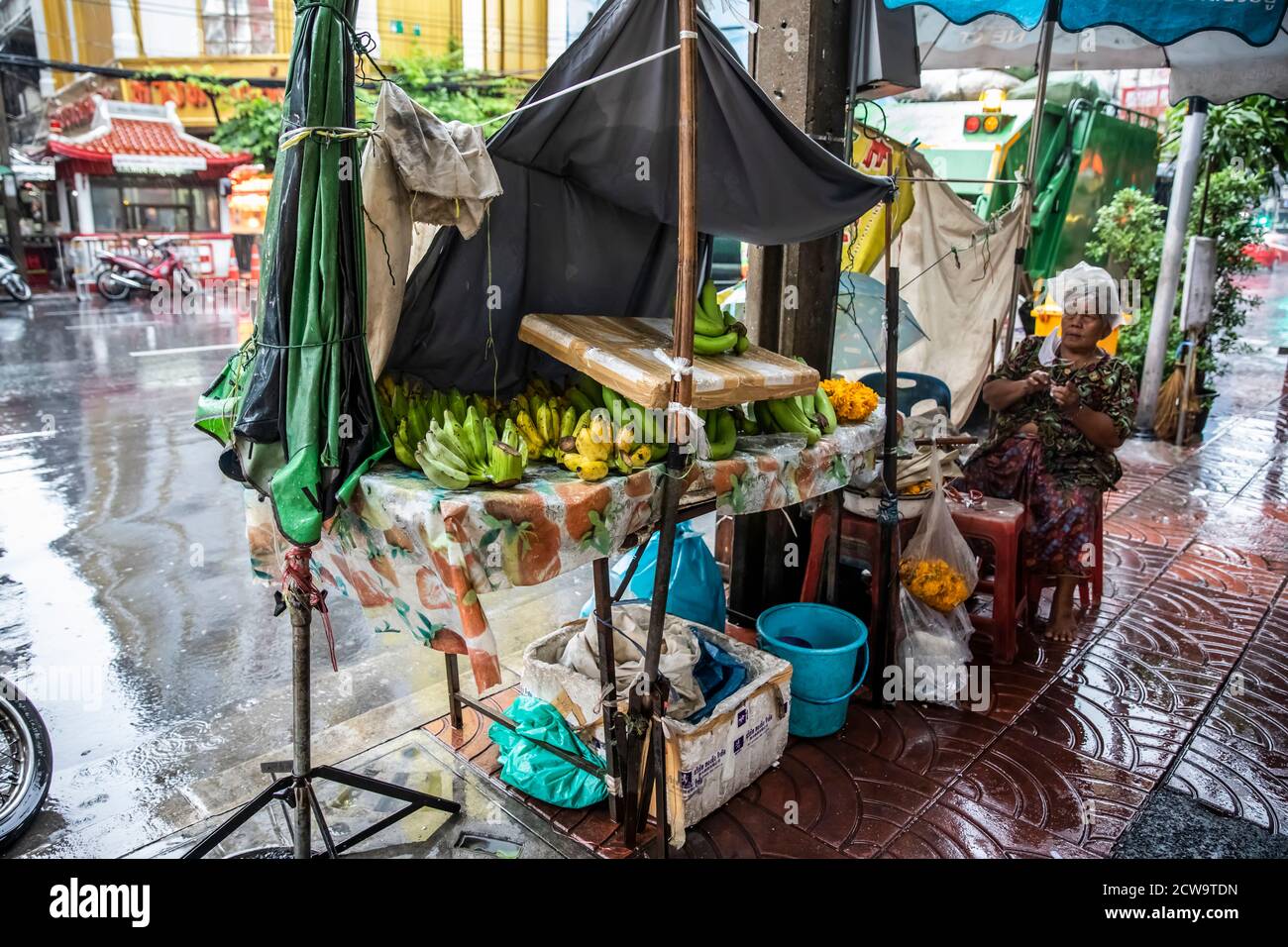 Ein Street-Food-Händler huddelt unter ihrem Regenschirm und trägt eine Plastiktüte auf ihrem Kopf, um während eines Regensturms in Bangkok, Chinatown, trocken zu bleiben. Stockfoto