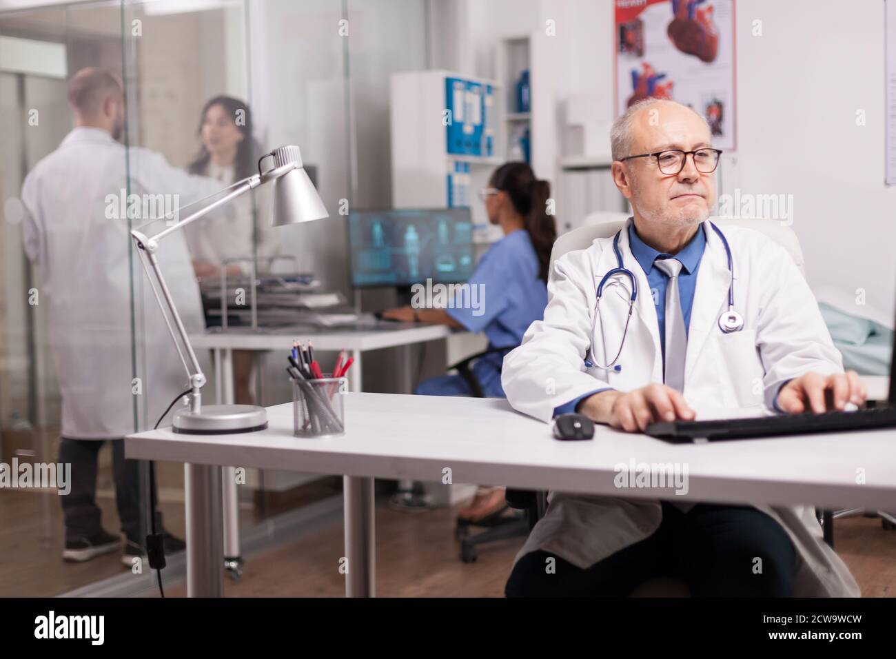 Fokussierter Arzt mit grauen Haaren Typisierung Wissenschaft Bericht über Computer im Krankenhaus Schrank und junge Sanitäter mit weißem Mantel helfen Patienten auf Klinik Korridor. Stockfoto