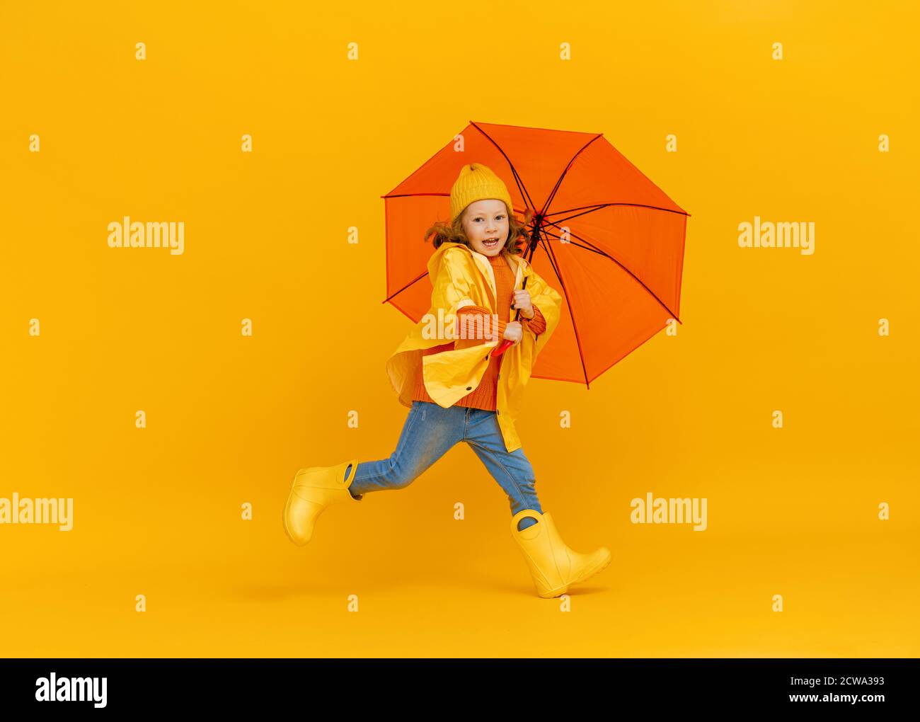 Glücklich emotionales Kind lachen und springen. Kind mit orangefarbenem Regenschirm auf farbigem gelbem Hintergrund. Stockfoto