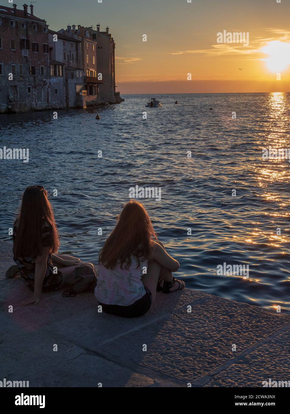 ROVINJ, KROATIEN - 06/23/2018: Zwei junge Frauen mit langen Haaren sitzen in der Nähe des Meeres und beobachten den Sonnenuntergang. Stockfoto