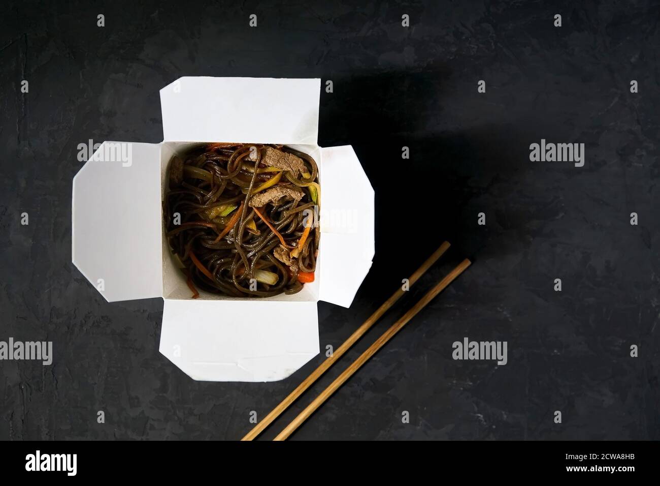 Asiatische Restaurant Essen Lieferung. Soba-Nudeln mit Fleisch, Gemüse und Sojasauce in weißer Take-Out-Papierbox auf schwarzem Hintergrund mit Essstäbchen, oben Stockfoto