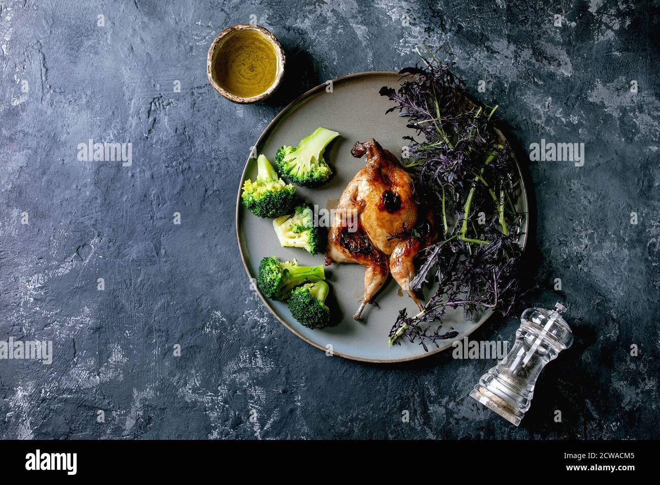 Gebratener gegrillter Schmetterlingsschwanz auf Keramikplatte mit garniergrünem Salat und Brokkoli, Pfeffermühle und Olivenöl über schwarzem Betonhintergrund. Flach Stockfoto