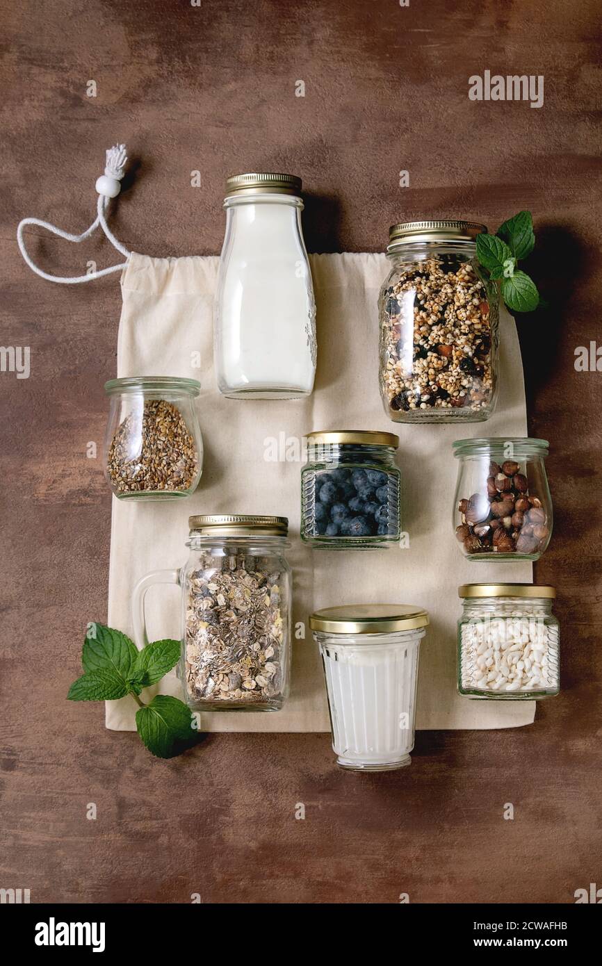 Hausgemachte knackige gebuffte Masergranola mit getrockneten Früchten und Nüssen, Joghurt, Milch, Müsli in verschiedenen Glasgefäßen auf Baumwolltasche. Brauner Hintergrund. Stockfoto