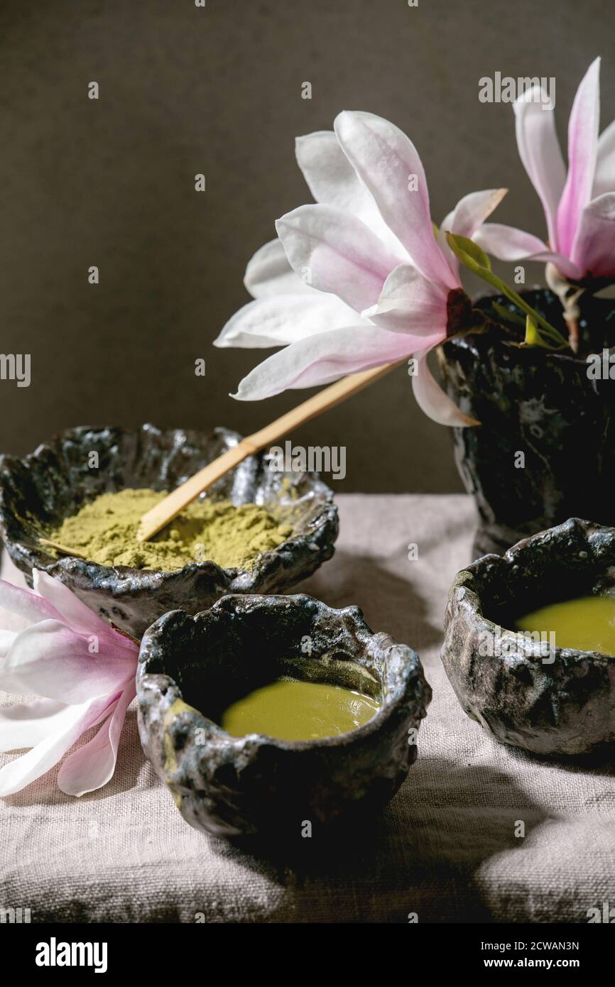 Japanischer Matcha grüner Tee in handwerklichen Keramikbechern mit Matcha-Pulver, Bambusbesen und rosa Magnolienblüten auf grauem Leinentischtuch. Stockfoto