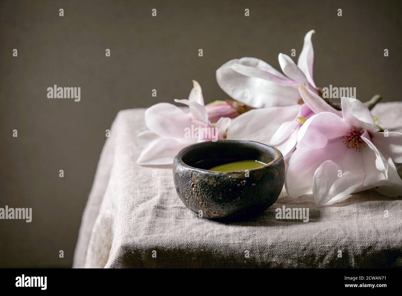 Japanischer Matcha-grüner Tee in Wabi Sabi Keramik-Tasse mit rosa Magnolienblüten auf grauem Leinentischtuch. Stockfoto