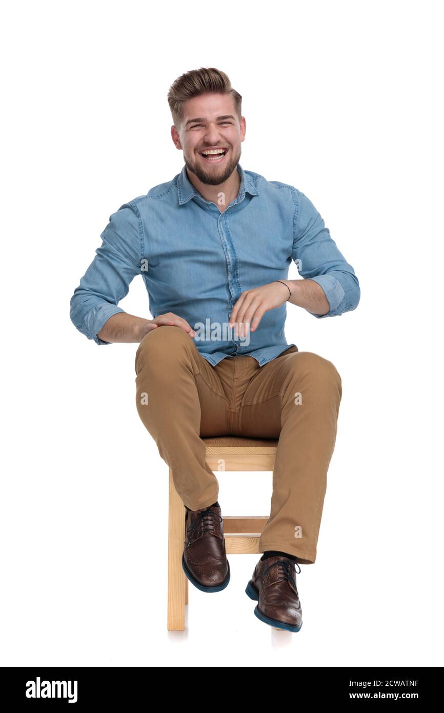 Fröhlicher Casual Mann trägt Denim-Shirt, hält die Hände auf Knie und lachend, sitzt isoliert auf weißem Hintergrund, voller Körper Stockfoto