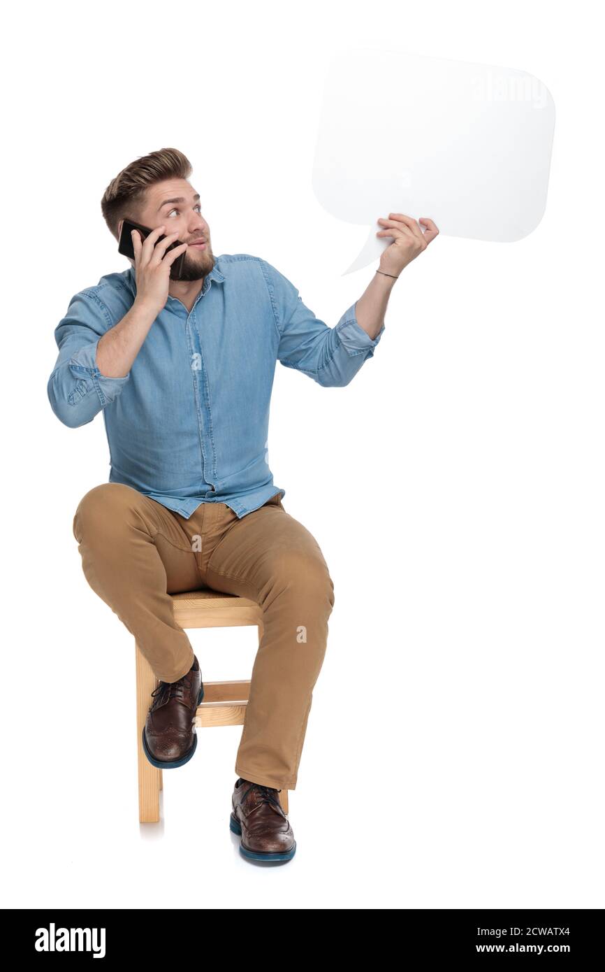 Verwirrt junger Mann am Telefon reden und halten Sprechblase, sitzen isoliert auf weißem Hintergrund, ganzer Körper Stockfoto