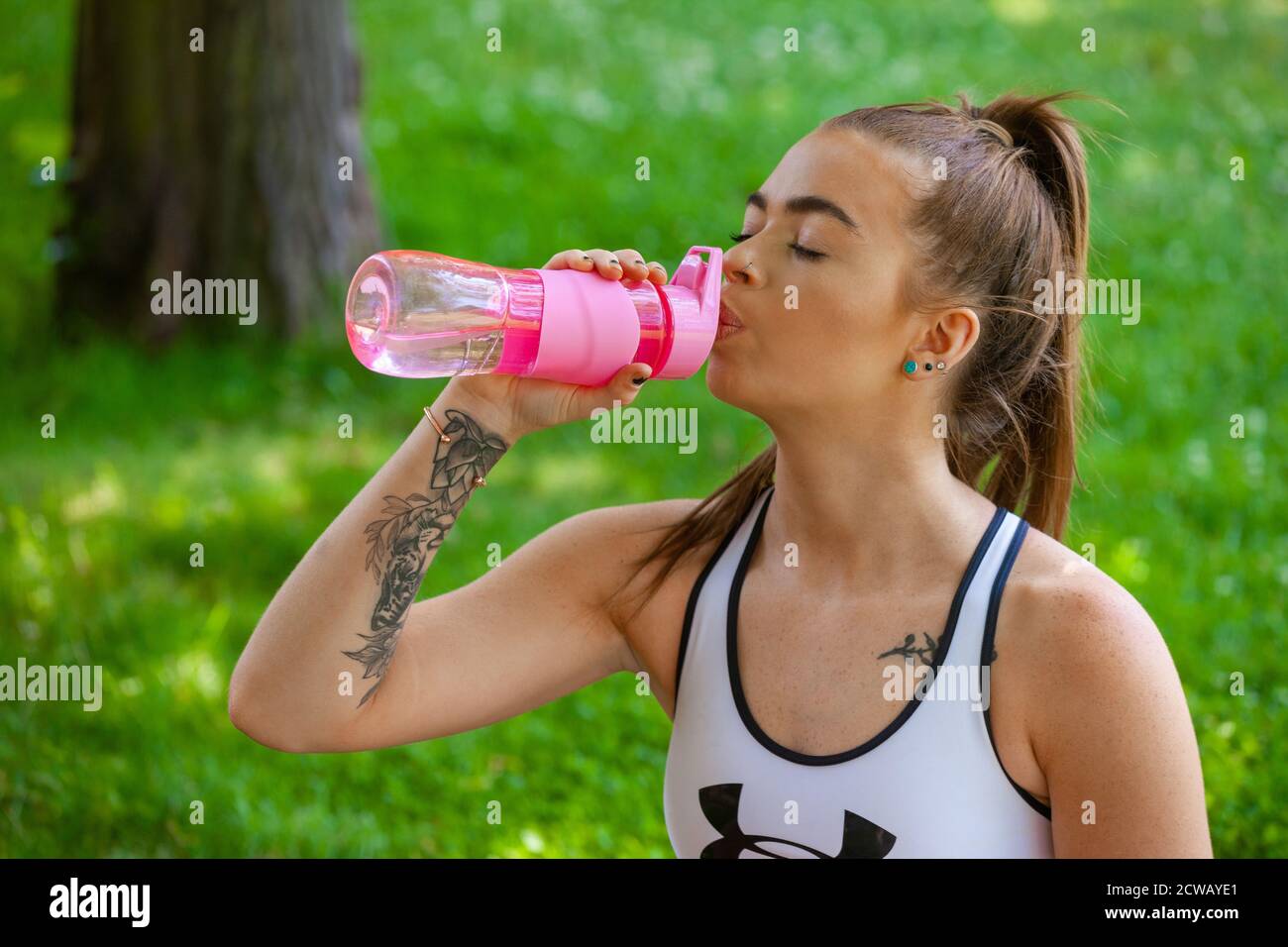 Eine junge Frau, die draußen Sportkleidung trägt und aus einer rosa Wasserflasche trinkt. Stockfoto