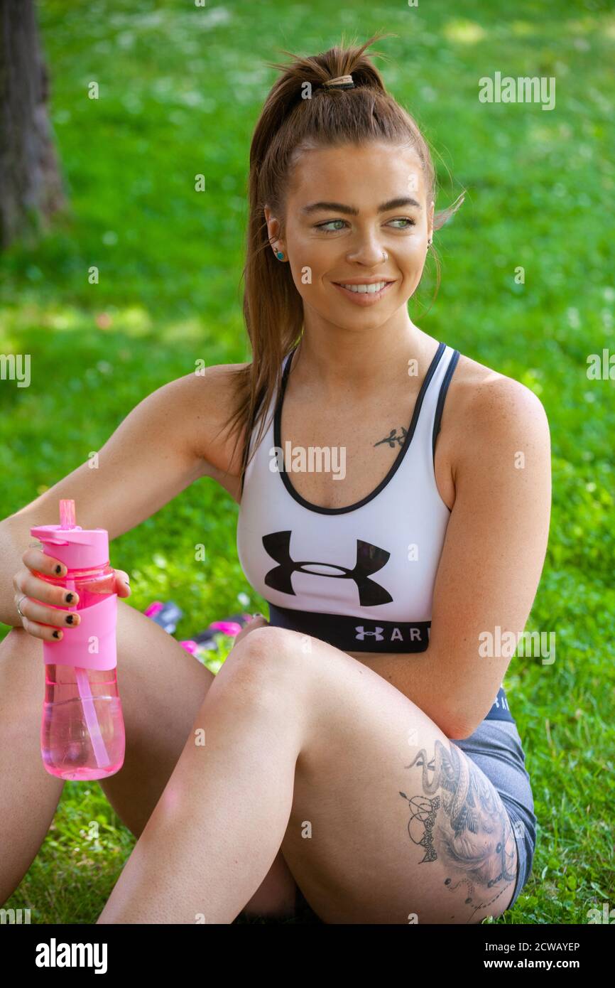 Eine junge Frau in Sportkleidung sitzt draußen auf Gras mit einer rosa Wasserflasche. Stockfoto