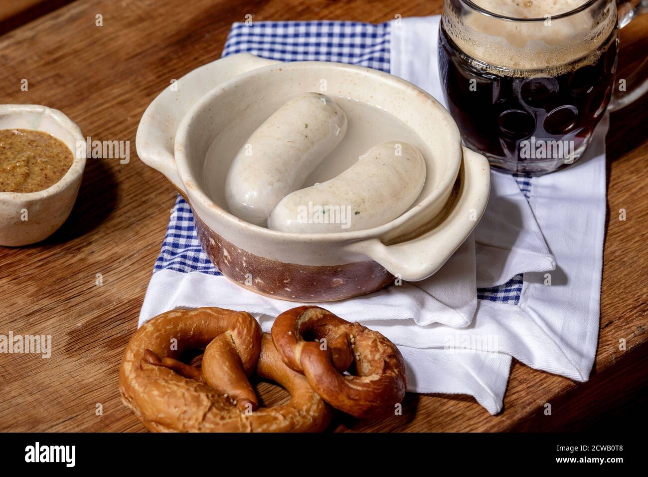 Münchner bayerische traditionelle Weißwürste in Keramikpfanne serviert mit deutschem süßen Senf, einem Becher dunklem Bier und Brezeln Brot auf weißer und blauer Serviette Stockfoto