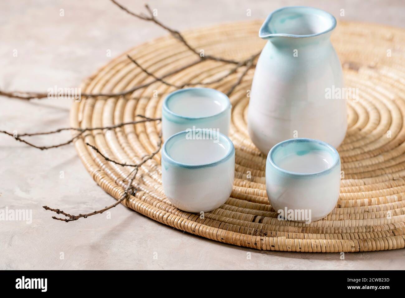 Sake Keramik Set für traditionellen japanischen Alkohol trinken Reiswein Sake, Krug und drei Tassen, stehen auf Strohserviette mit trockenen Zweigen über beige Stockfoto