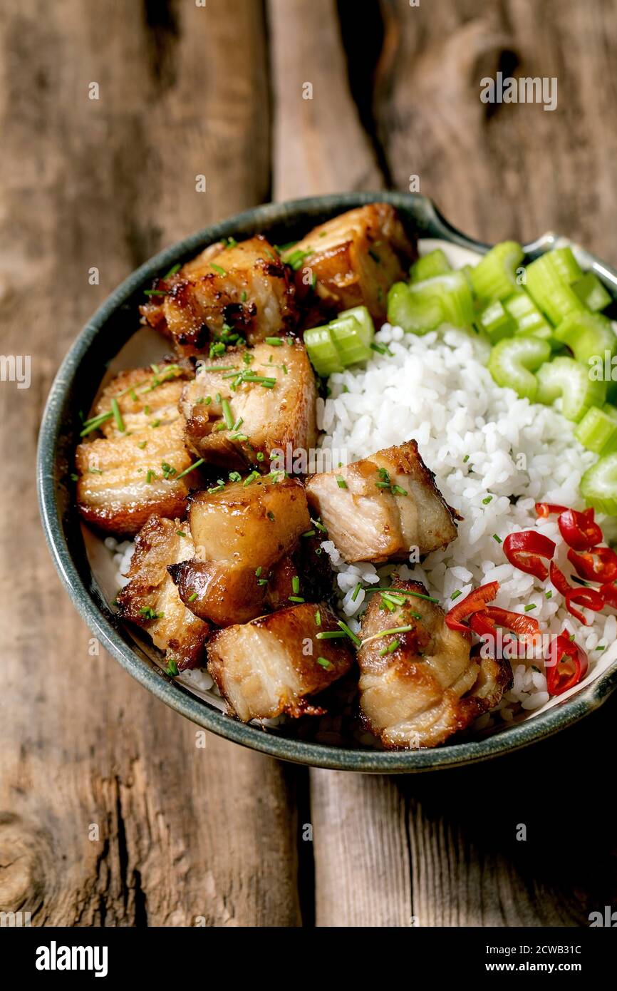 Gegrillter frittierter Schweinebauch in einer Schüssel mit Reis, Sellerie, Chili und Frühlingszwiebeln auf altem Holzhintergrund. Stockfoto