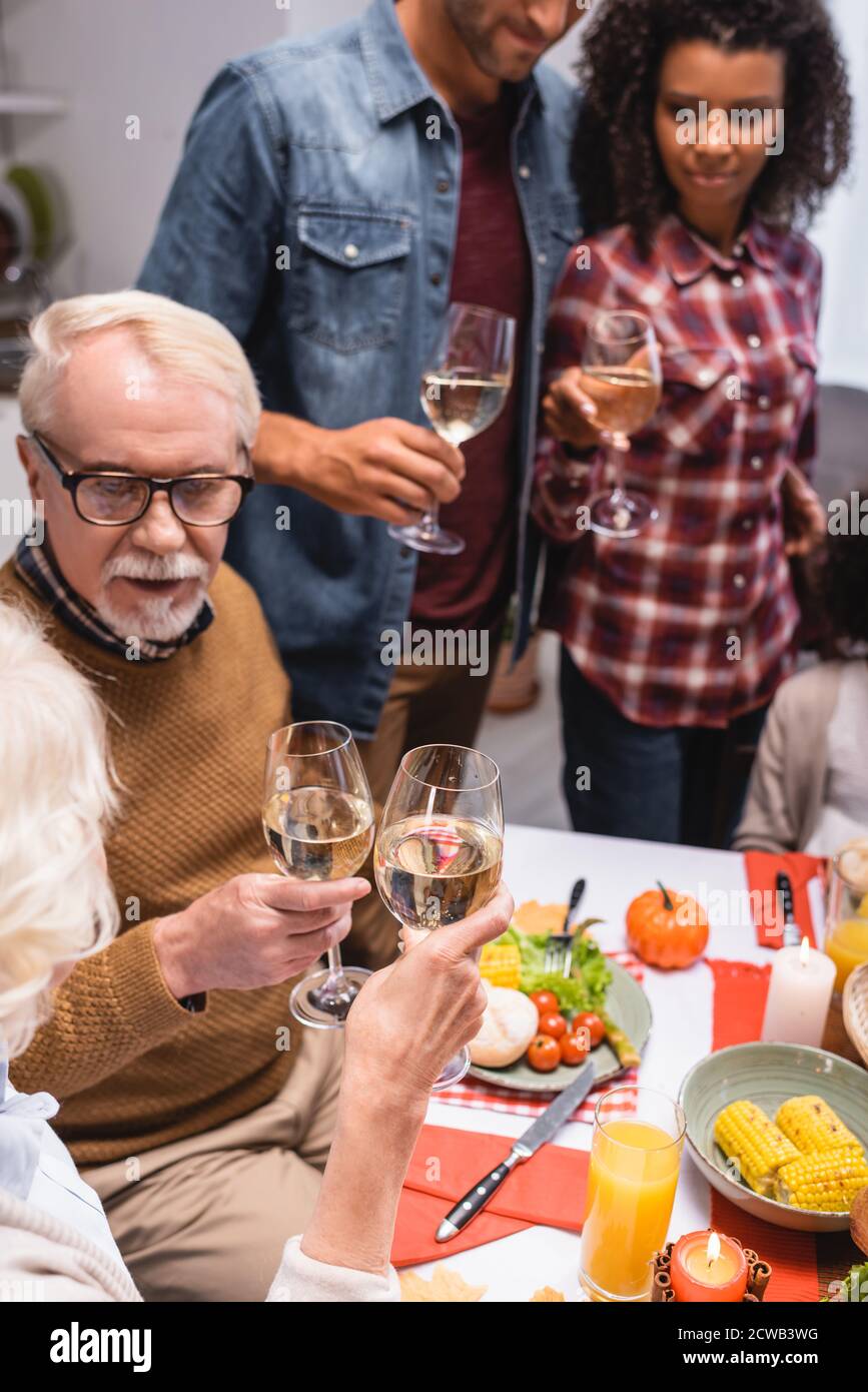 Selektiver Fokus auf ältere Frau, die während des Weinanklickens mit Mann anklickend thanksgiving-Feier mit multiethnischen Familie Stockfoto