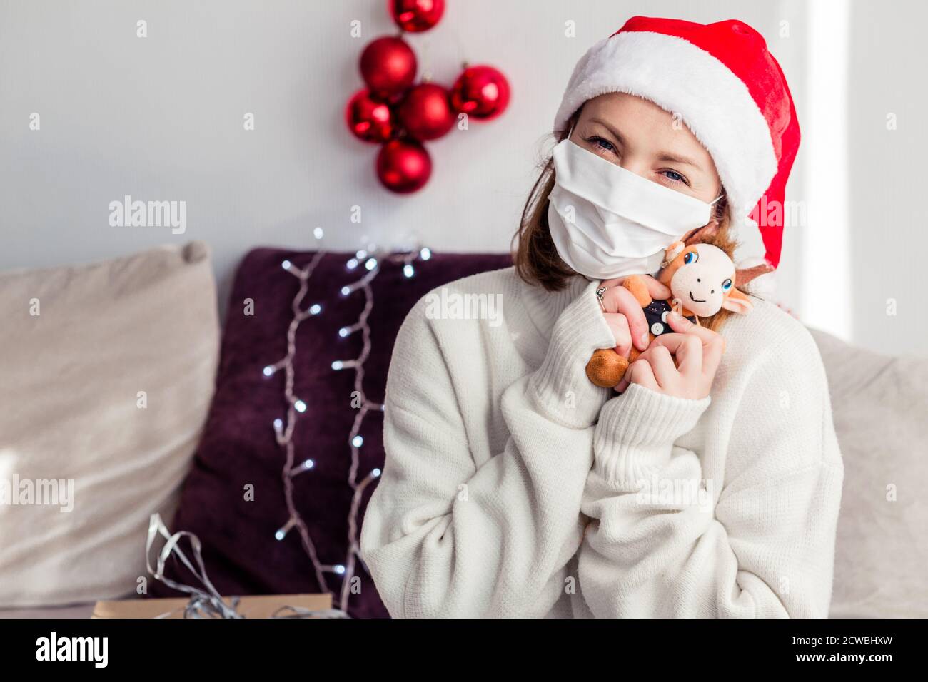 Eine junge Frau in einem weißen Pullover, Weihnachtsmann Hut, medizinische Maske schaut in die Kamera und umarmt einen weichen Spielzeug-Stier. Stockfoto