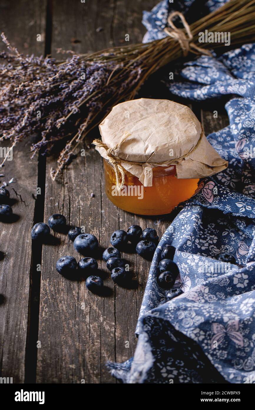 Glas Glas von flüssigen Honig mit Wabenstruktur innen, frische Blaubeeren und Bündel trockener Lavendel über alten Holztisch mit blauen Textilien Rag. Dunklen rustikalen Stockfoto