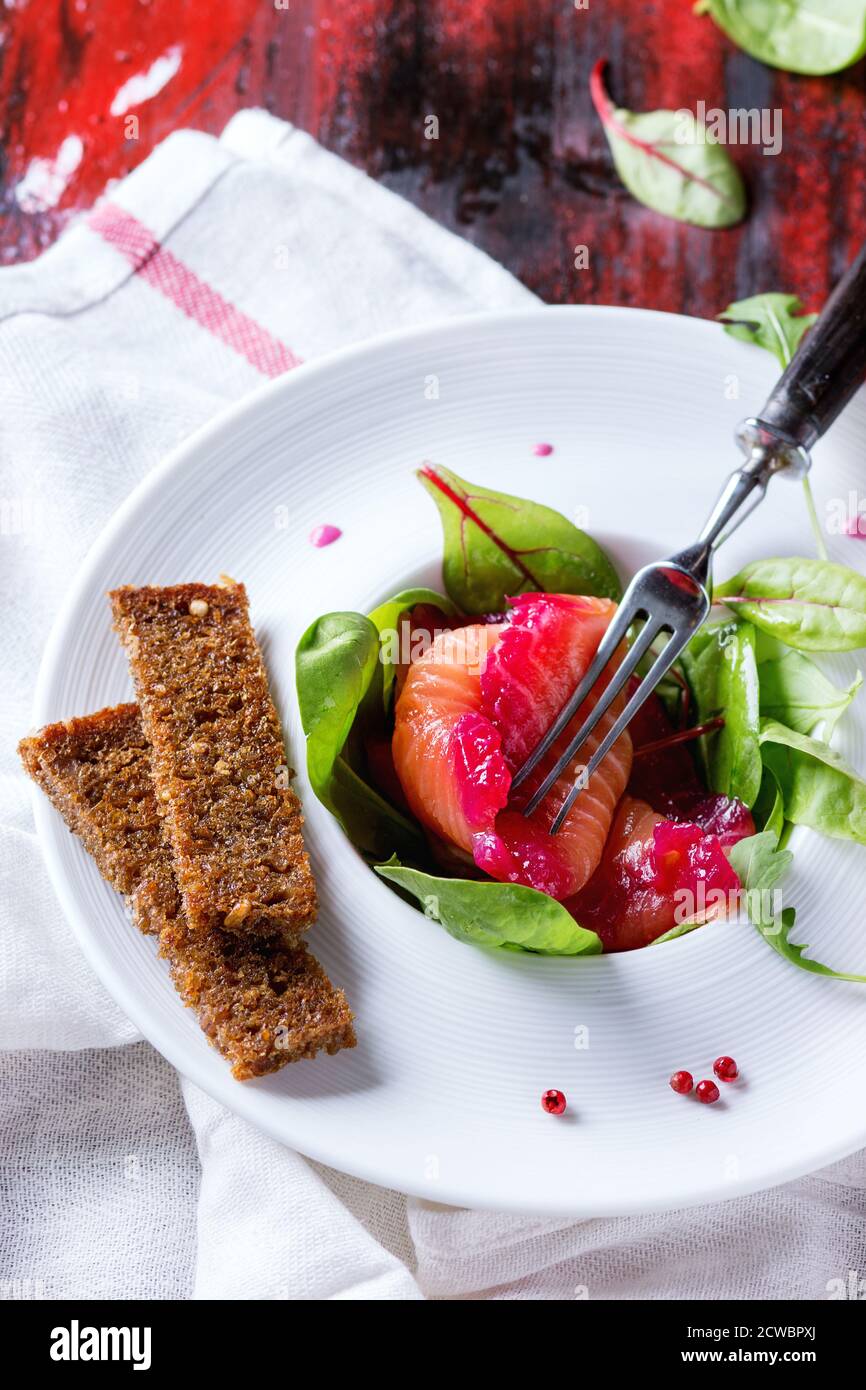 Weiße Keramikplatte mit Lachsfilet in Scheiben, gesalzen mit Rote Bete Saft, serviert mit Vollkorntoasts, Salatblättern und Rote Bete Sauce über rotem Woo Stockfoto