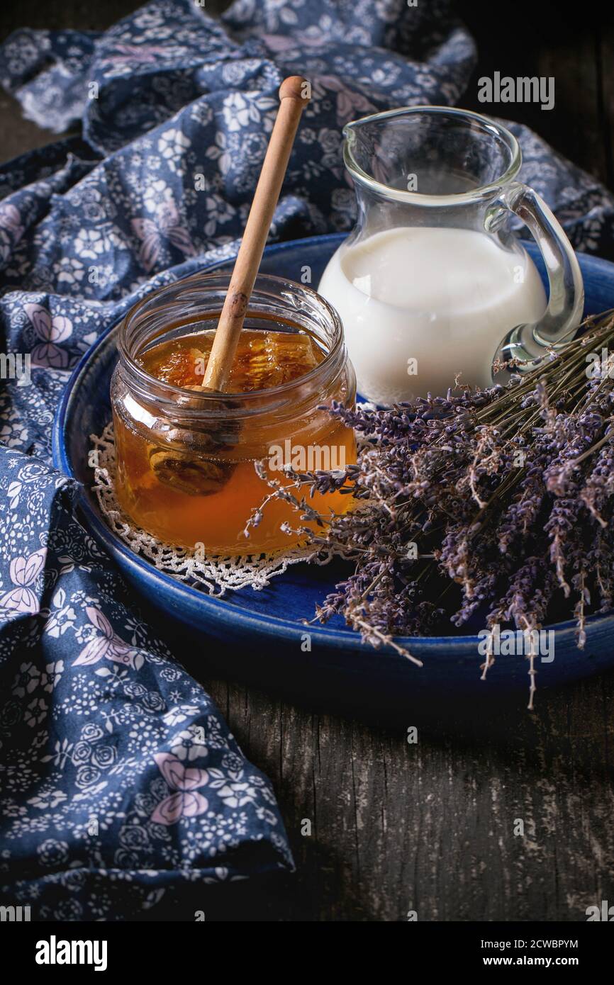 Offenes Glas mit flüssigem Honig mit Honigwabe und Honig-Dipper innen, Glaskrug mit Milch und Bund von trockenem Lavendel in blauer Keramikplatte über Holz s Stockfoto