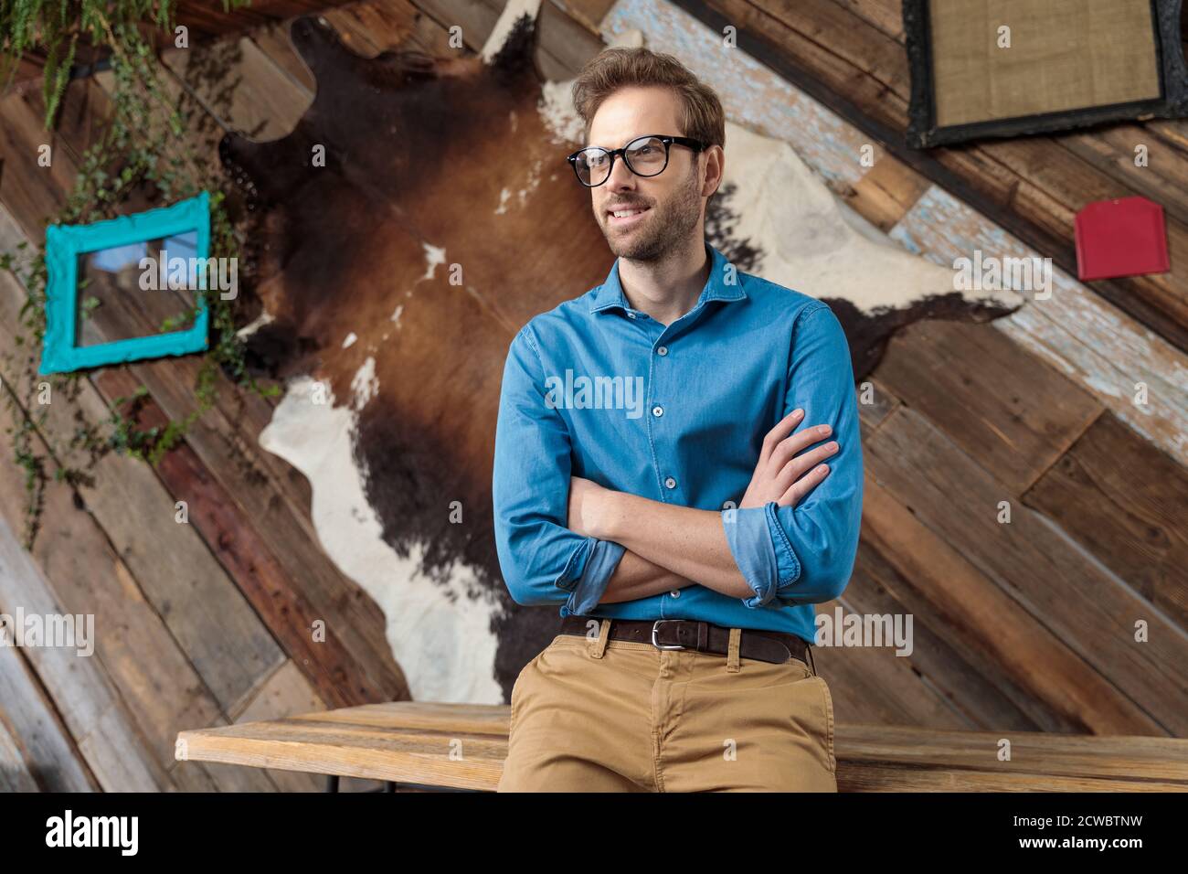 Hoffnungsvolles Model schaut weg, lächelnd mit gekreuzten Armen, während es blaues Hemd und Brille trägt und auf einem Schreibtisch auf Coffeeshop Hintergrund sitzt Stockfoto